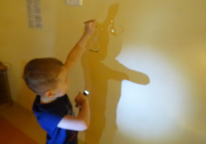Chłopiec trzyma w ręku okulary na które świeci latarką, tworząc cień na ścienie.